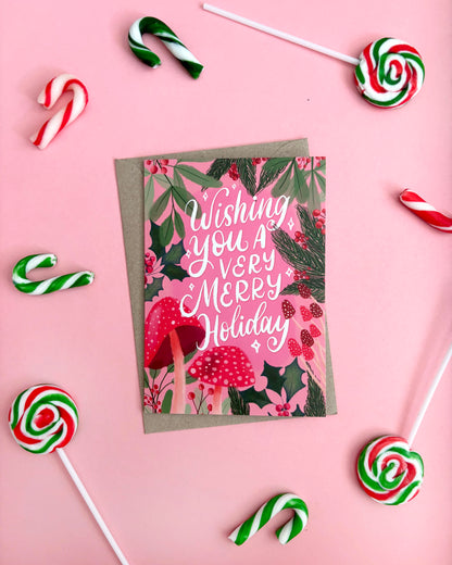 A festive greeting card with a kraft envelope, featuring the text 'Wishing you a VERY Merry Holiday' in white script over a Christmas-themed illustration of red-and-white mushrooms, berries, and green foliage. The card is styled in a flat-lay on a pink background, surrounded by candy canes and lollipops.