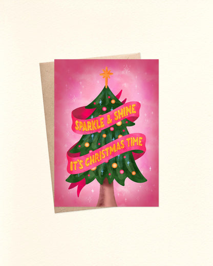 A6 Christmas card featuring a whimsical watercolor Christmas tree in deep green with yellow, pink, and orange baubles, topped with a yellow star. A bright pink ribbon swirls around the tree with yellow hand-lettered text reading “Sparkle and Shine, It’s Christmas Time.” The background is pink with tiny white stars. The card is shown laid flat on top of a brown kraft envelope.