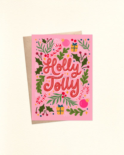 A6 Christmas card with a pale pink background and bold red hand-lettered text reading “Holly Jolly” in the centre. Surrounding the text are festive illustrations including holly leaves, red berries, candy canes, wrapped presents, baubles, and other foliage. The card is laid flat with a brown kraft envelope beneath it.