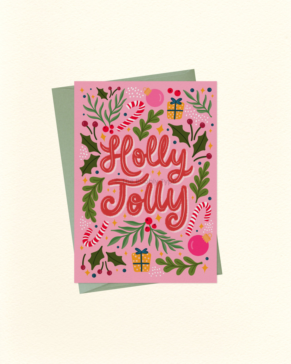 A pink Christmas greeting card with the words 'Holly Jolly' in red script, surrounded by holly leaves, candy canes, ornaments, and gift boxes, shown with a muted green envelope on a cream background.