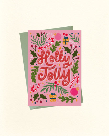 A pink Christmas greeting card with the words 'Holly Jolly' in red script, surrounded by holly leaves, candy canes, ornaments, and gift boxes, shown with a muted green envelope on a cream background.