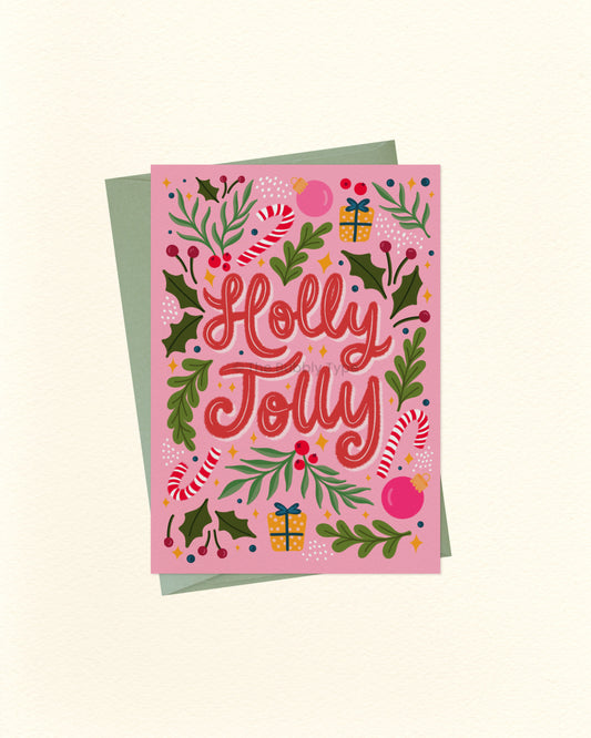 A pink Christmas greeting card with the words 'Holly Jolly' in red script, surrounded by holly leaves, candy canes, ornaments, and gift boxes, shown with a muted green envelope on a cream background.