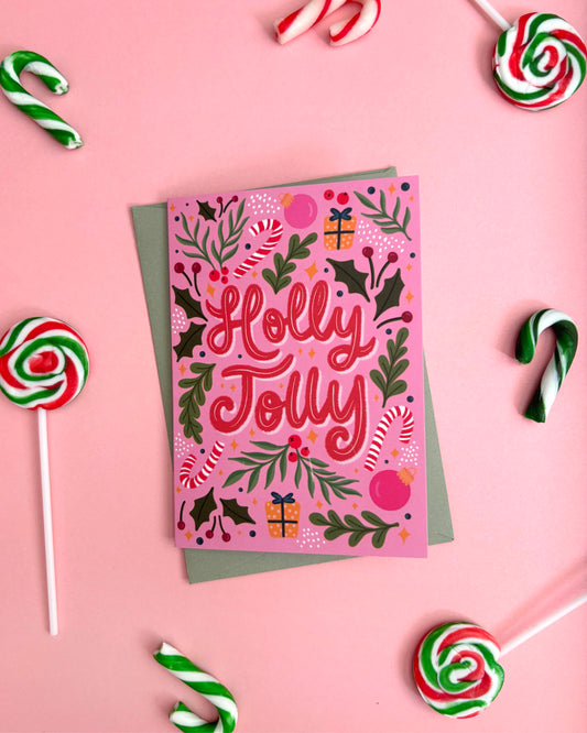 A pink Christmas greeting card with 'Holly Jolly' in red script, surrounded by festive illustrations of holly leaves, candy canes, ornaments, and gift boxes. The card is layered over a muted green envelope and photographed from above on a pink background, with candy canes and lollipops arranged around it.