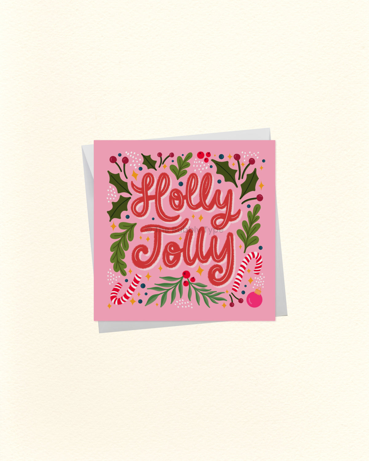 A square pink greeting card with the words 'Holly Jolly' in red script, surrounded by green holly leaves, red berries, and candy canes, shown on a light cream background with a soft shadow.