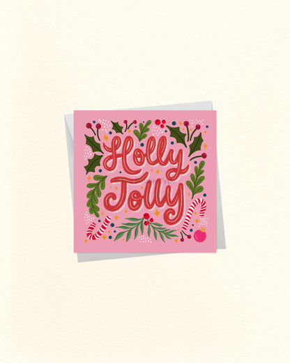 A square pink greeting card with the words 'Holly Jolly' in red script, surrounded by green holly leaves, red berries, and candy canes, shown on a light cream background with a soft shadow.