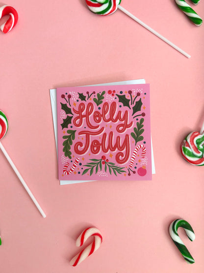 A square pink Christmas greeting card with 'Holly Jolly' written in red script, surrounded by holly leaves, berries, and candy canes, styled on a pink background with candy lollipops.