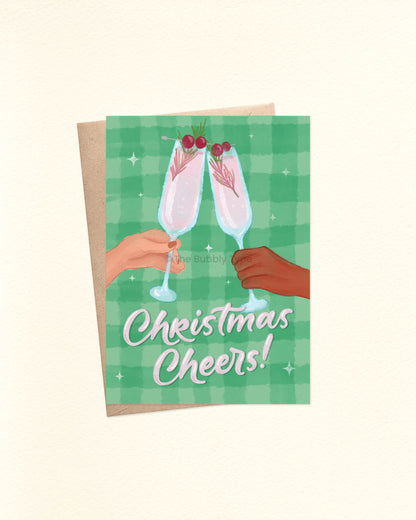 A6 Christmas card front design featuring an illustration of two hands—one pale and one brown—clinking champagne flutes filled with pink bubbly and sprigs of pine leaves with cherries. Below the hands, in pale pink hand lettering, the message reads “Christmas Cheers.” The background is a green watercolour checkered pattern with white stars. A kraft brown envelope peeks out from underneath the card.