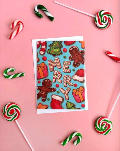 A greeting card with a festive Christmas design featuring gingerbread men, candy canes, gifts, and the word 'MERRY' on a light blue background, styled on a pink surface with candy canes, lollipops, and a striped stick around it.