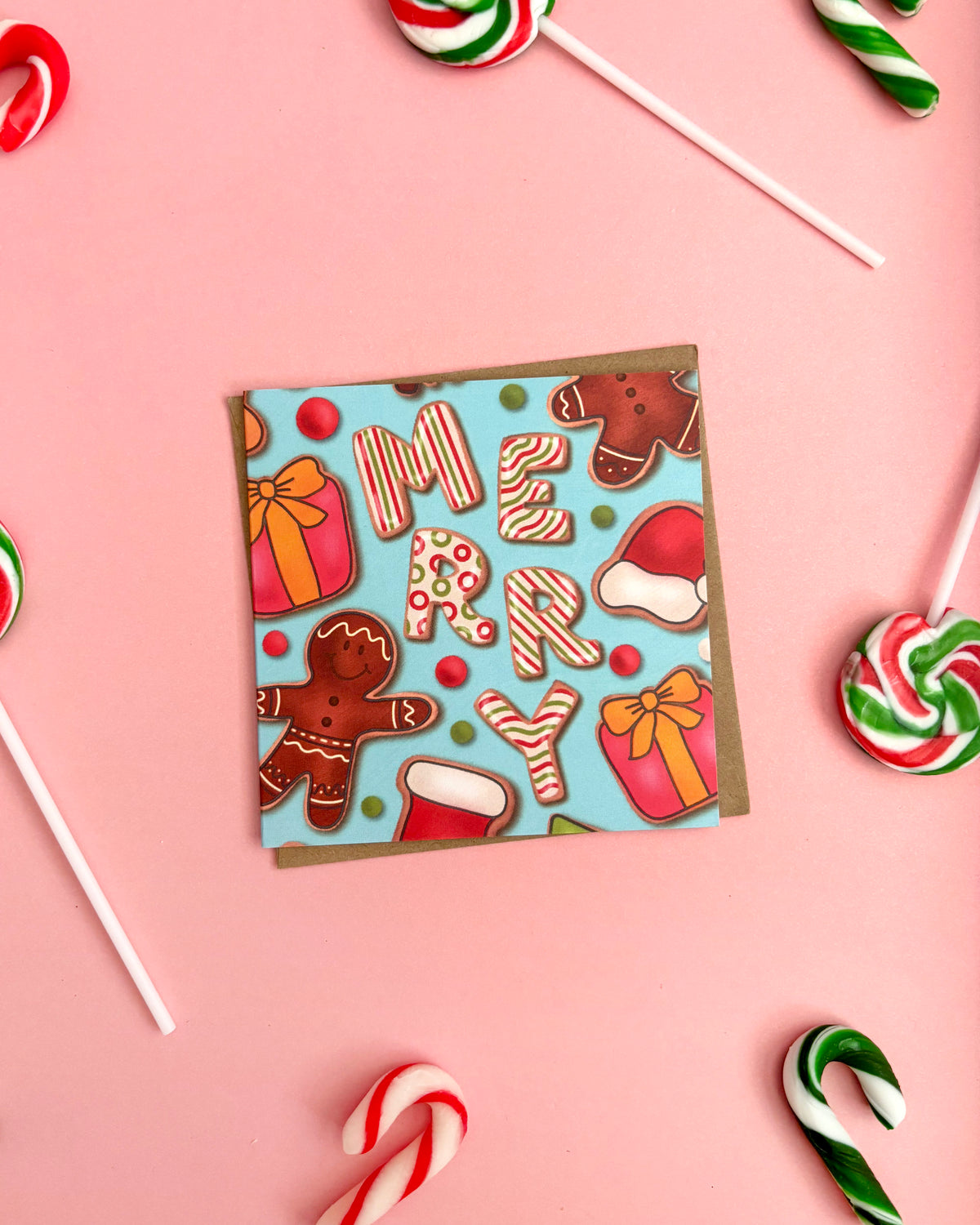 A square paper card with a festive Christmas design featuring gingerbread men, candy canes, Santa hats, and the word 'MERRY' on a light blue background, styled on a pink surface with Christmas candy props.