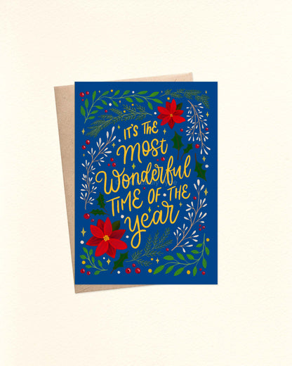 A6 Christmas greeting card with deep blue background, yellow lettering reading ‘It’s the most wonderful time of the year’, surrounded by holly and festive foliage, displayed on a brown kraft envelope.