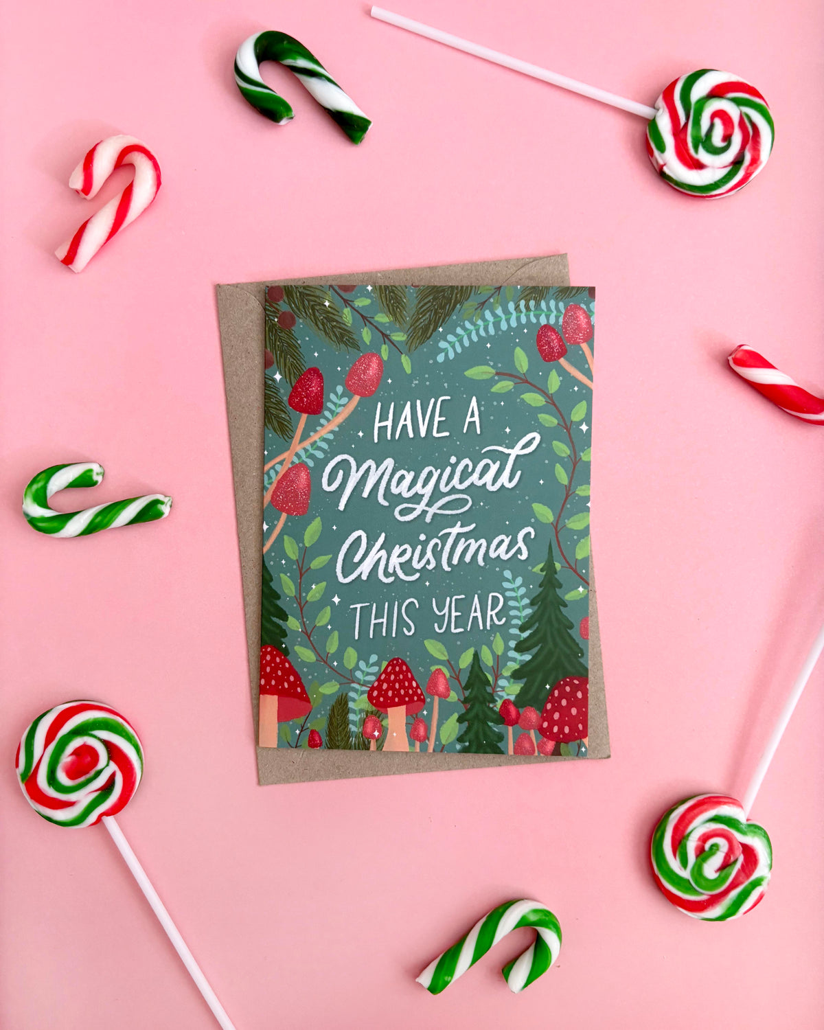 A Christmas greeting card with a kraft envelope, styled in a flat-lay on a pink background with candy canes and lollipops. The card features a festive forest illustration with the text 'HAVE A Magical Christmas THIS YEAR'.