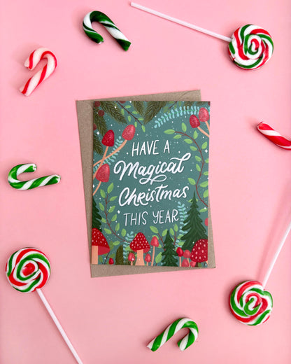 A Christmas greeting card with a kraft envelope, styled in a flat-lay on a pink background with candy canes and lollipops. The card features a festive forest illustration with the text 'HAVE A Magical Christmas THIS YEAR'.
