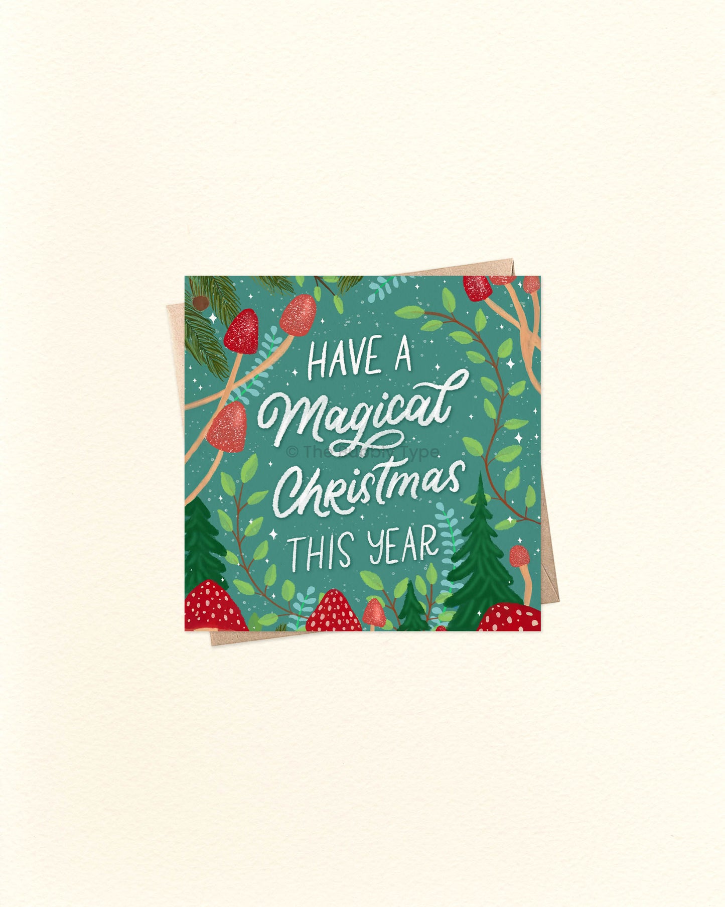 Square Christmas card with illustrated red mushrooms and pine foliage, featuring the message ‘Have a Magical Christmas This Year’, placed over a kraft brown envelope. Buy unique Christmas cards in Australia with whimsical designs.