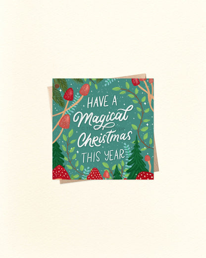 Square Christmas card with illustrated red mushrooms and pine foliage, featuring the message ‘Have a Magical Christmas This Year’, placed over a kraft brown envelope. Buy unique Christmas cards in Australia with whimsical designs.