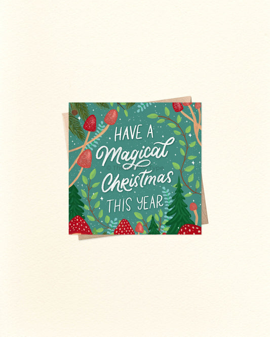 Square Christmas card with illustrated red mushrooms and pine foliage, featuring the message ‘Have a Magical Christmas This Year’, placed over a kraft brown envelope. Buy unique Christmas cards in Australia with whimsical designs.