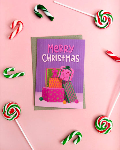 A Christmas greeting card with a purple background and 'Merry Christmas' text, decorated with colourful gift boxes. The card is placed on a pink background with candy canes and lollipops around it.