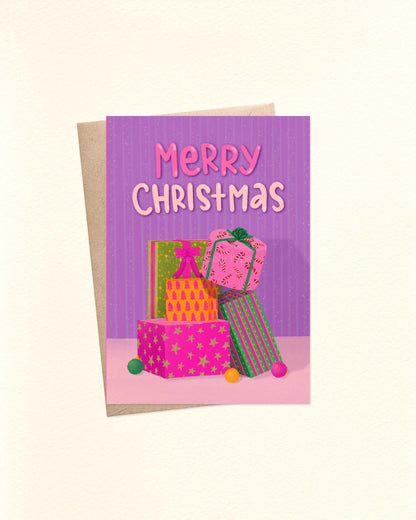 A6 Christmas card featuring a light purple background, illustration of Christmas gift stack, and hand-lettered ‘Merry Christmas’ at the top, laid on a kraft brown envelope.