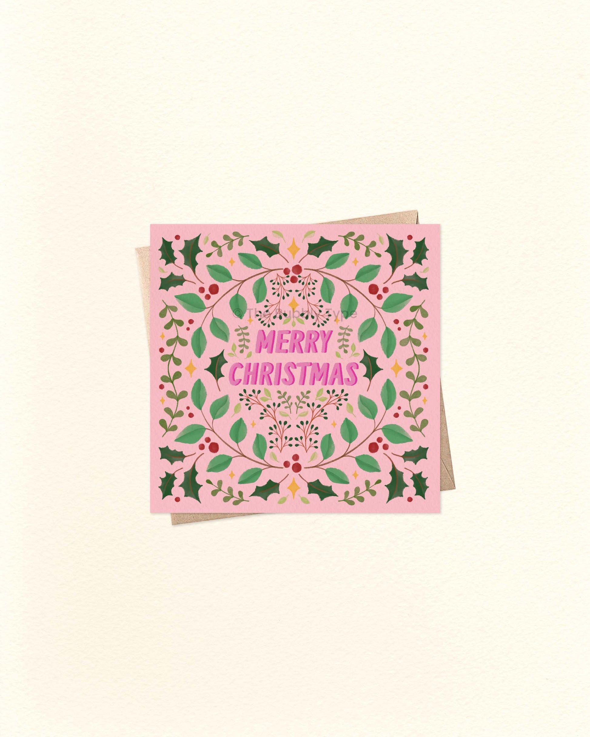 Square pink Christmas greeting card with festive foliage border and bold ‘Merry Christmas’ lettering in the centre, shown on a brown kraft envelope.