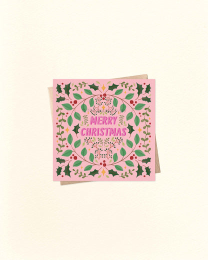Square pink Christmas greeting card with festive foliage border and bold ‘Merry Christmas’ lettering in the centre, shown on a brown kraft envelope.