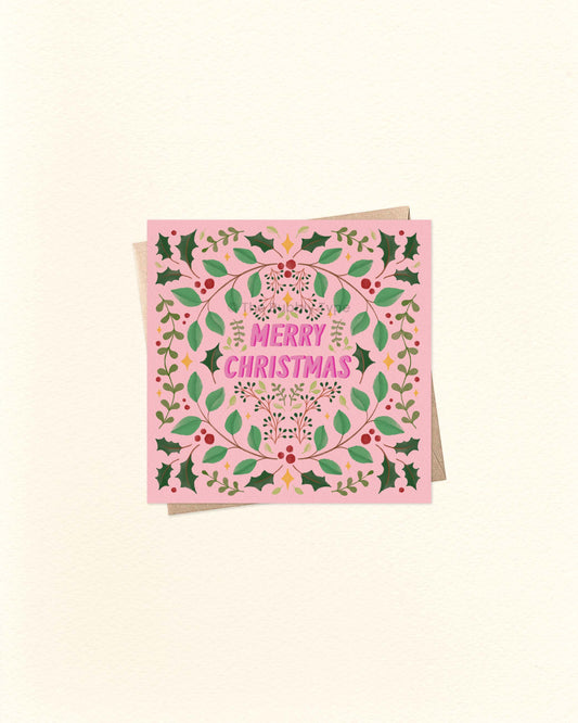 Square pink Christmas greeting card with festive foliage border and bold ‘Merry Christmas’ lettering in the centre, shown on a brown kraft envelope.
