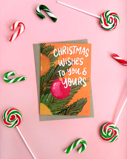 A Christmas greeting card with the text 'Christmas Wishes to You & Yours' in white script, decorated with green pine branches, a pink ornament on an orange background. The card is placed on a kraft envelope and photographed from above on a pink background, surrounded by candy canes and lollipops.