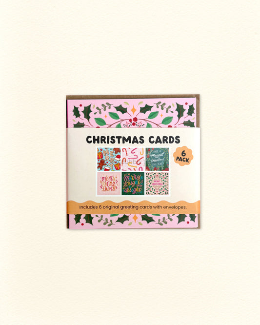 A boxed set of Christmas greeting cards shown against a plain light beige background. The package has a pink and green holly border and reads 'CHRISTMAS CARDS' with '6 pieces' and 'Includes 6 original greeting cards with envelopes' on the front.