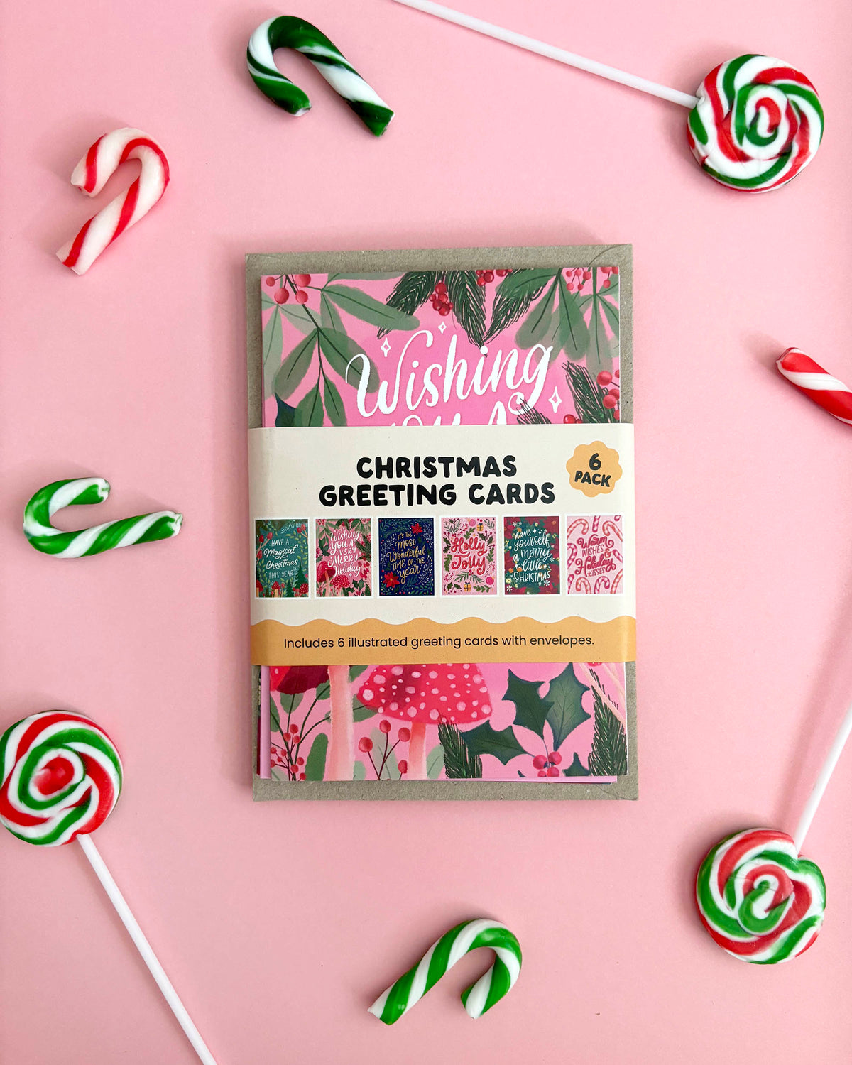 A flat-lay of a Christmas greeting card pack on a pink background, surrounded by candy canes and lollipops. The greeting cards feature hand illustrated festive designs with pink, green, and red colours.
