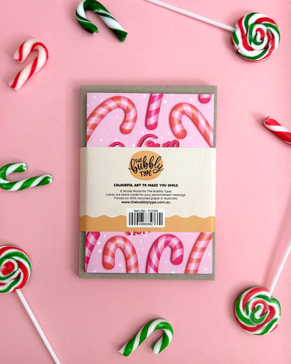 The back of a Christmas-themed greeting card pack styled in a flat-lay with candy canes and lollipops on a pink background. 
