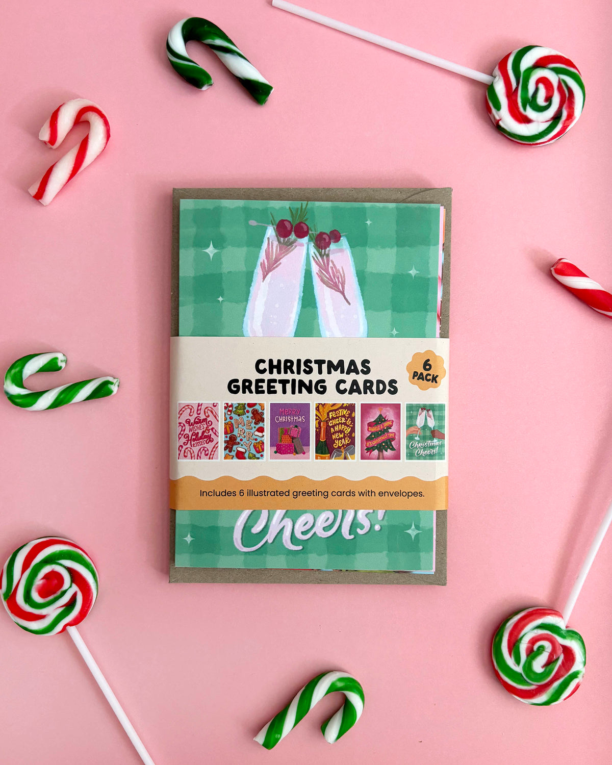 A 6-piece Christmas greeting card set with brown kraft envelopes, styled on a pink background with candy canes and lollipops. The card packaging features a green plaid pattern card peeking out behind a belly band with the text 'Christmas Greeting Cards' and '6 Pack'.