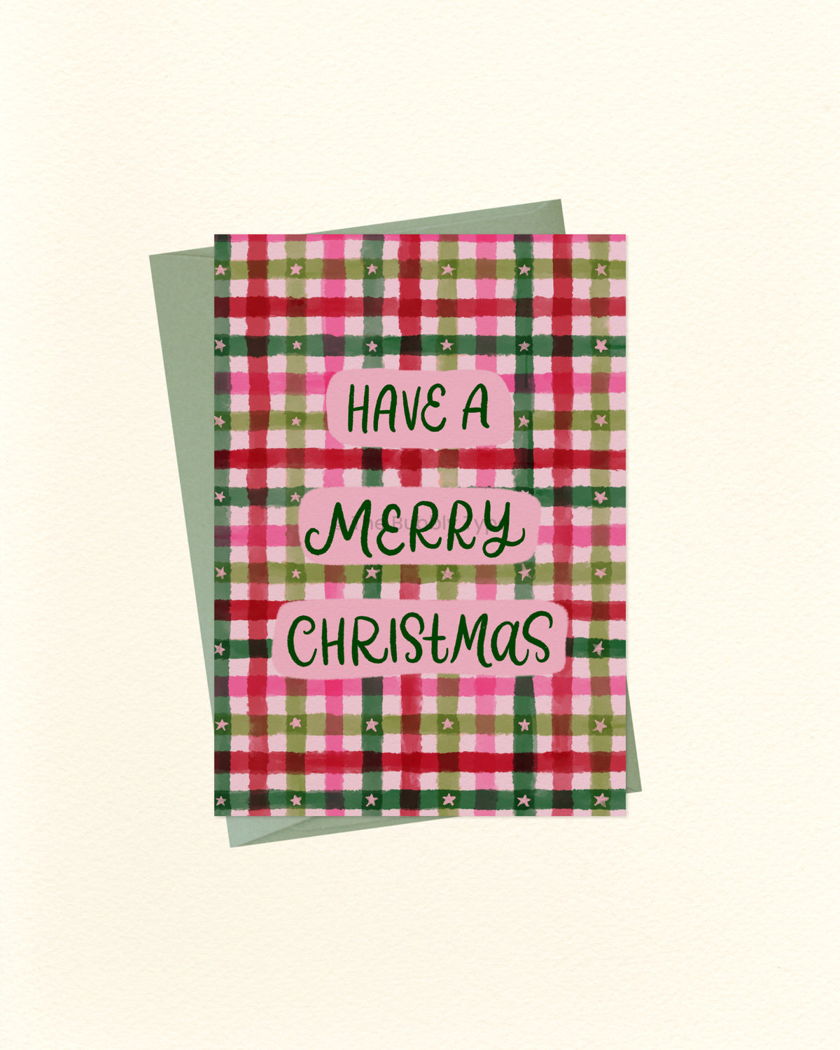 A Christmas greeting card with a pink, green, and red plaid pattern, featuring the text 'HAVE A MERRY CHRISTMAS' in black script on pink labels. A sage green envelope is placed behind the card on a light cream background.