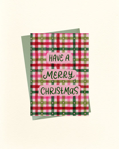 A Christmas greeting card with a pink, green, and red plaid pattern, featuring the text 'HAVE A MERRY CHRISTMAS' in black script on pink labels. A sage green envelope is placed behind the card on a light cream background.