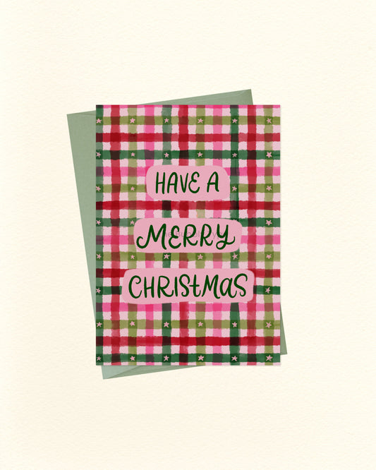 A Christmas greeting card with a pink, green, and red plaid pattern, featuring the text 'HAVE A MERRY CHRISTMAS' in black script on pink labels. A sage green envelope is placed behind the card on a light cream background.