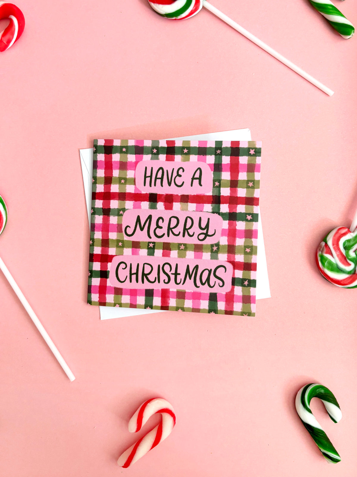 A square Christmas greeting card with a pink, green, and red plaid pattern and the words 'HAVE A MERRY CHRISTMAS' in black script, styled on a pink background with candy canes and lollipops.