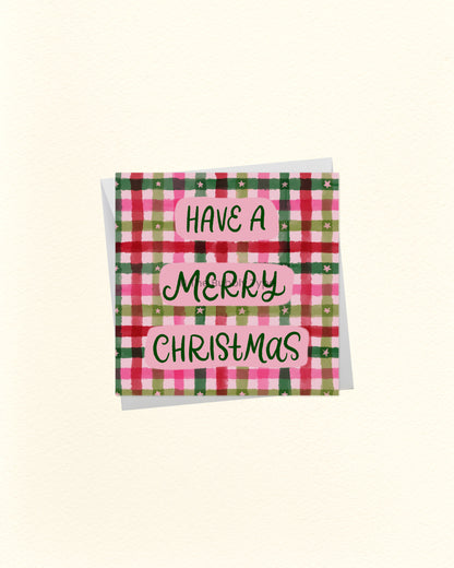 A square Christmas greeting card with a pink, green, and red plaid pattern and the words 'HAVE A MERRY CHRISTMAS' in black script on pink labels, shown on a cream background.