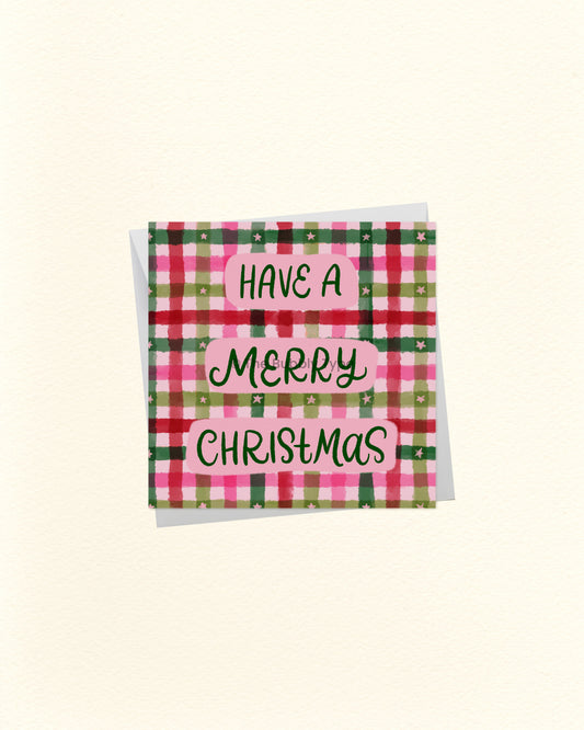 A square Christmas greeting card with a pink, green, and red plaid pattern and the words 'HAVE A MERRY CHRISTMAS' in black script on pink labels, shown on a cream background.