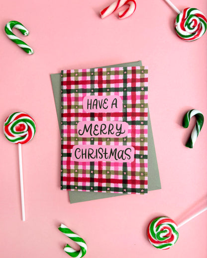 A square Christmas greeting card with a pink, green, and black plaid pattern and the text 'HAVE A MERRY CHRISTMAS' in white, styled on a pink background with candy canes and lollipops.