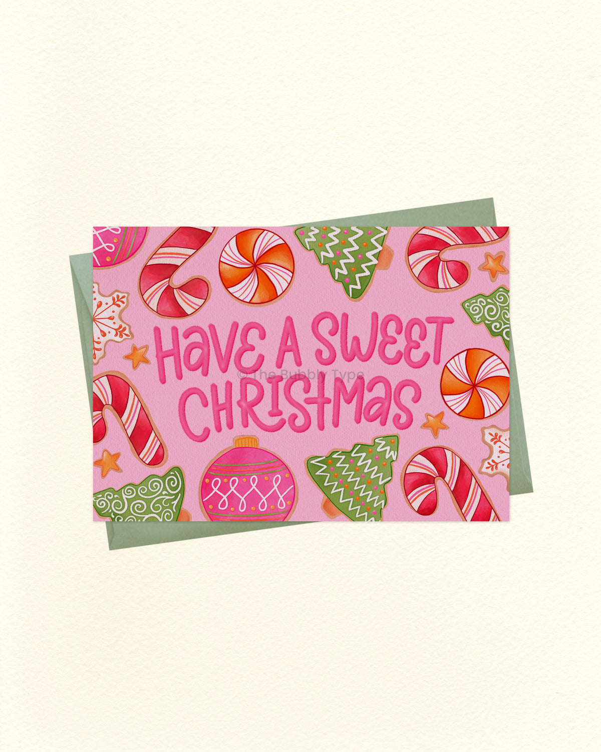 A square Christmas greeting card with a pink background and the phrase “Have a Sweet Christmas” in pink lettering, surrounded by illustrated candy canes, ornaments, stars, and green trees, shown with a muted green envelope on a light cream background.