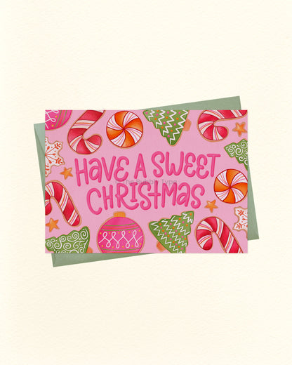 A square Christmas greeting card with a pink background and the phrase “Have a Sweet Christmas” in pink lettering, surrounded by illustrated candy canes, ornaments, stars, and green trees, shown with a muted green envelope on a light cream background.