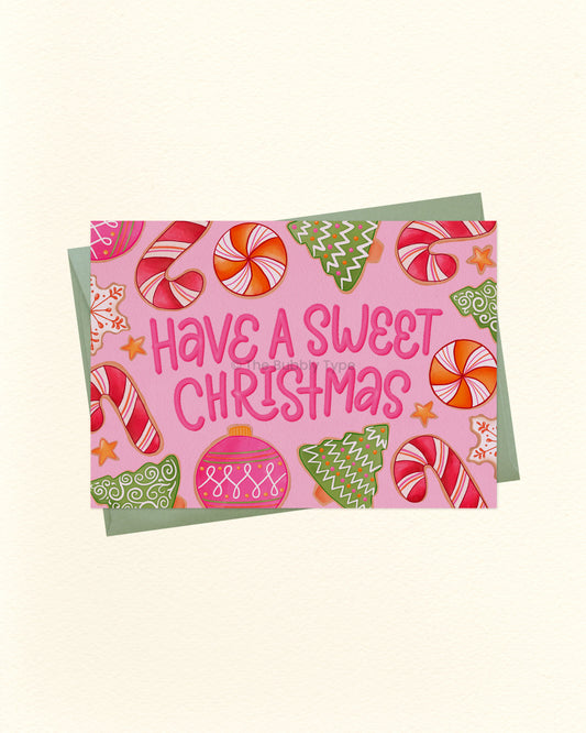 A square Christmas greeting card with a pink background and the phrase “Have a Sweet Christmas” in pink lettering, surrounded by illustrated candy canes, ornaments, stars, and green trees, shown with a muted green envelope on a light cream background.
