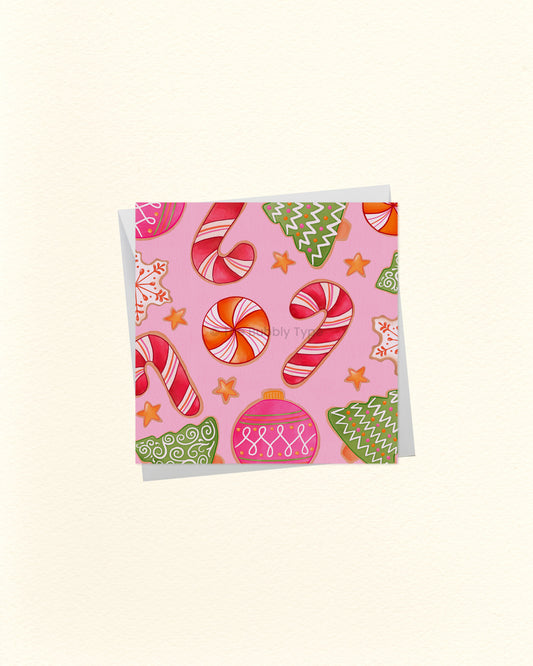 A square greeting card with a pink background and a festive holiday pattern featuring candy canes, ornaments, stars, and gingerbread cookies, shown on a light cream background.