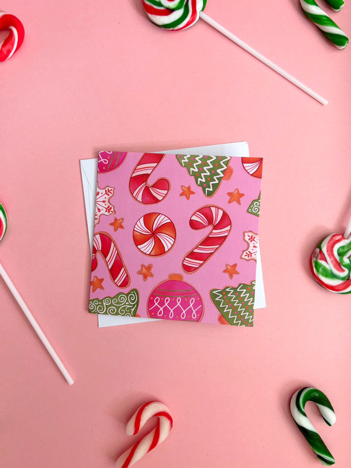 A square gift card with a pink background and a festive candy cane and Christmas tree pattern, displayed on top of a white envelope against a solid pink background with candy lollipops scattered around.