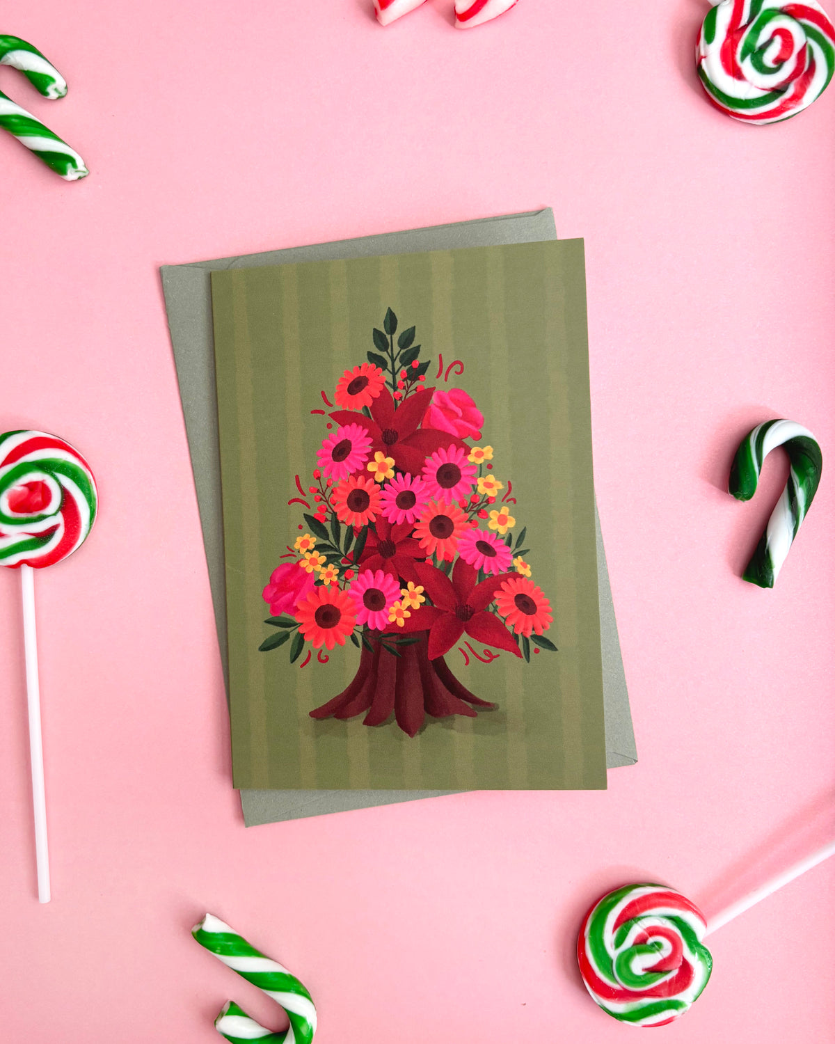 A greeting card featuring a colourful floral Christmas tree illustration, displayed on a pink background with candy cane props.