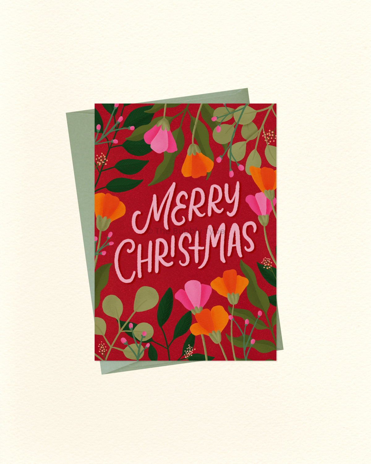 A square Christmas greeting card with a red floral design and the words “Merry Christmas” in white script, shown with a muted green envelope behind it on a light cream background.