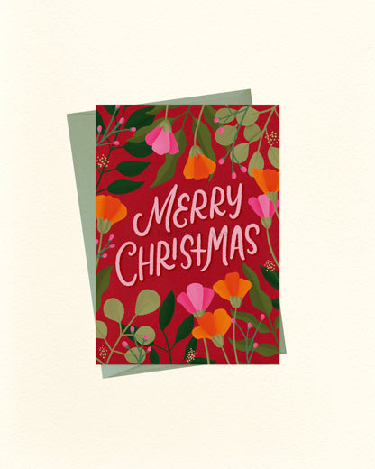 A square Christmas greeting card with a red floral design and the words “Merry Christmas” in white script, shown with a muted green envelope behind it on a light cream background.