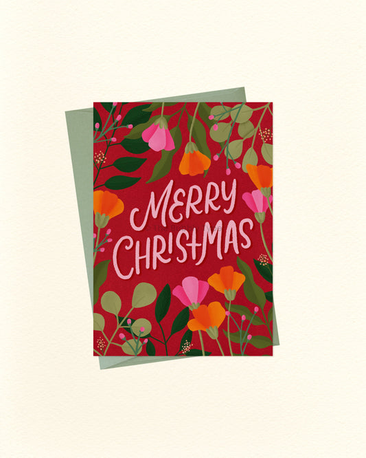 A square Christmas greeting card with a red floral design and the words “Merry Christmas” in white script, shown with a muted green envelope behind it on a light cream background.