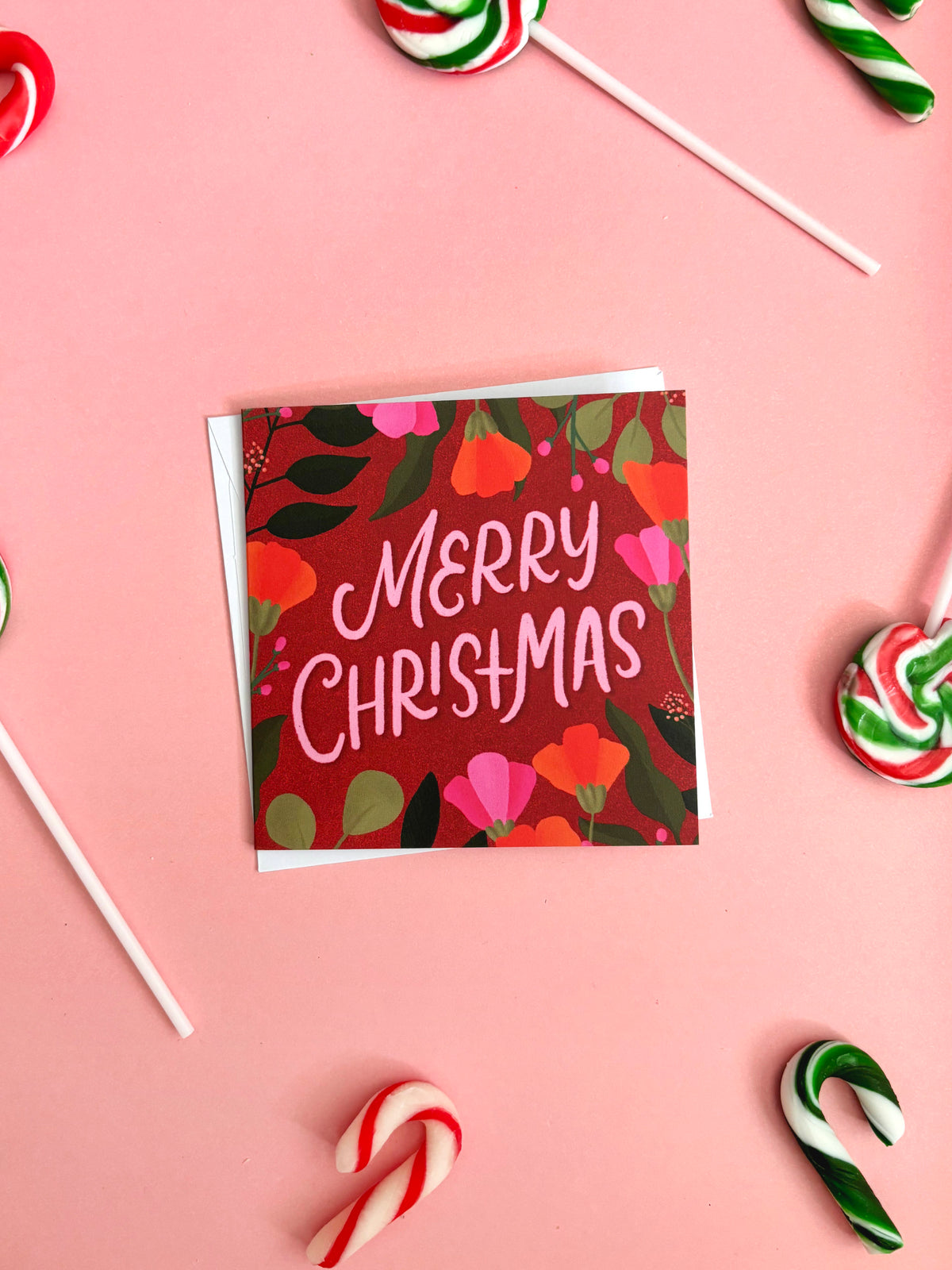 A square Christmas greeting card with a floral pattern and the words 'Merry Christmas' in white script, placed on a pink background with candy canes and lollipops around it.