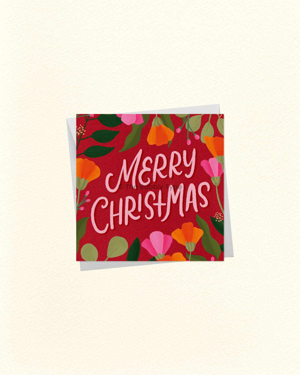 A square Christmas greeting card with a red background and colourful floral border, featuring the words “Merry Christmas” in white script, shown with a white envelope behind it on a light cream background.