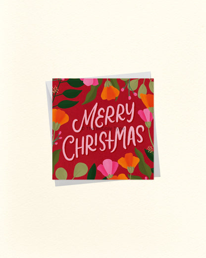 A square Christmas greeting card with a red background and colourful floral border, featuring the words “Merry Christmas” in white script, shown with a white envelope behind it on a light cream background.