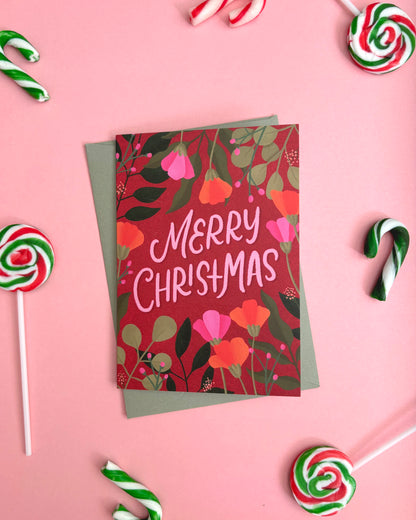 A Christmas greeting card with a red floral pattern and the words 'MERRY CHRISTMAS' in white script, layered over a muted green envelope. The card is photographed from above on a pink background, surrounded by candy canes and lollipops.