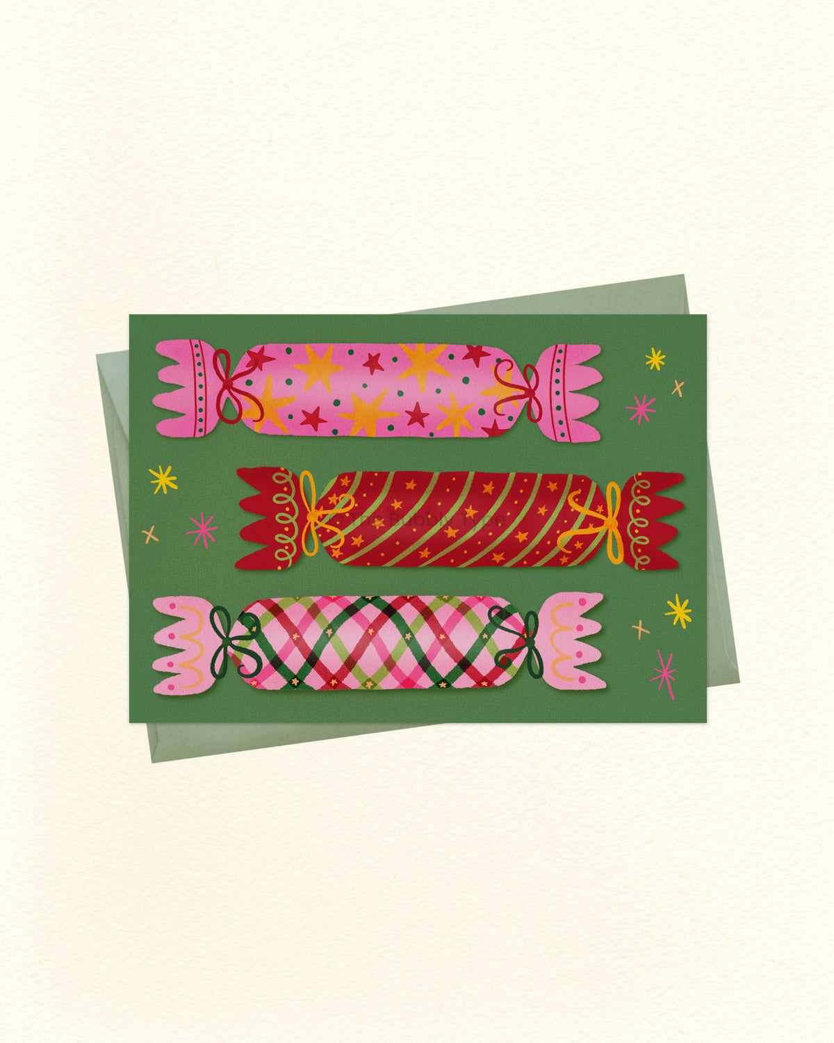 A square art print or card featuring three stylized Christmas crackers in pink, red, and green plaid patterns on a dark green background, with small yellow star accents. A light green envelope is partially visible behind the card on a warm off-white background.